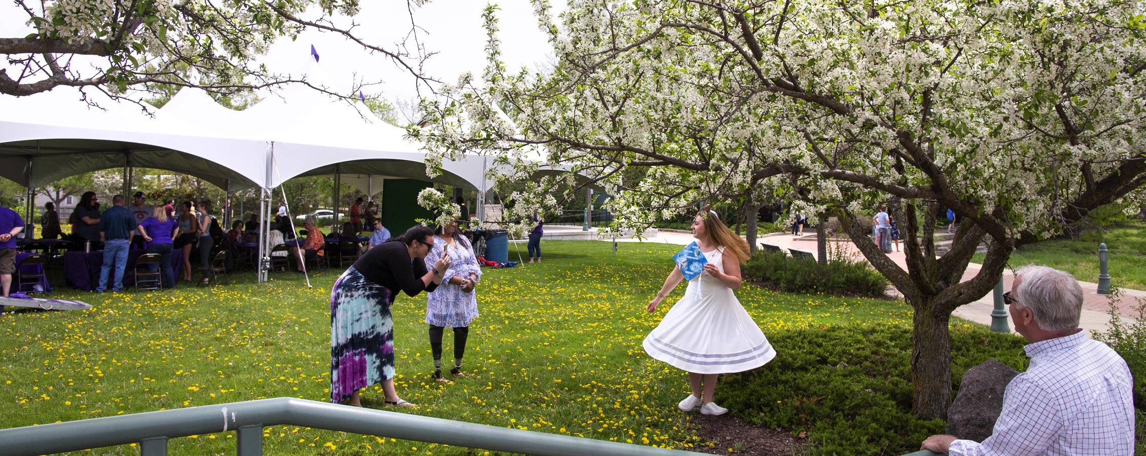 A person stands amongst flowering trees in a white dress and twirls as another person takes their photo.